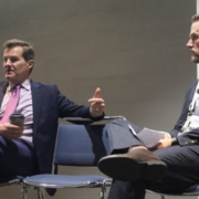 Two men in suits sitting on chairs and one is pointing at the camera.